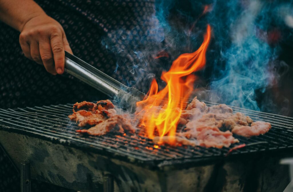 Close-up of grilling meat with vibrant flames and smoky aura, showcasing outdoor cooking.