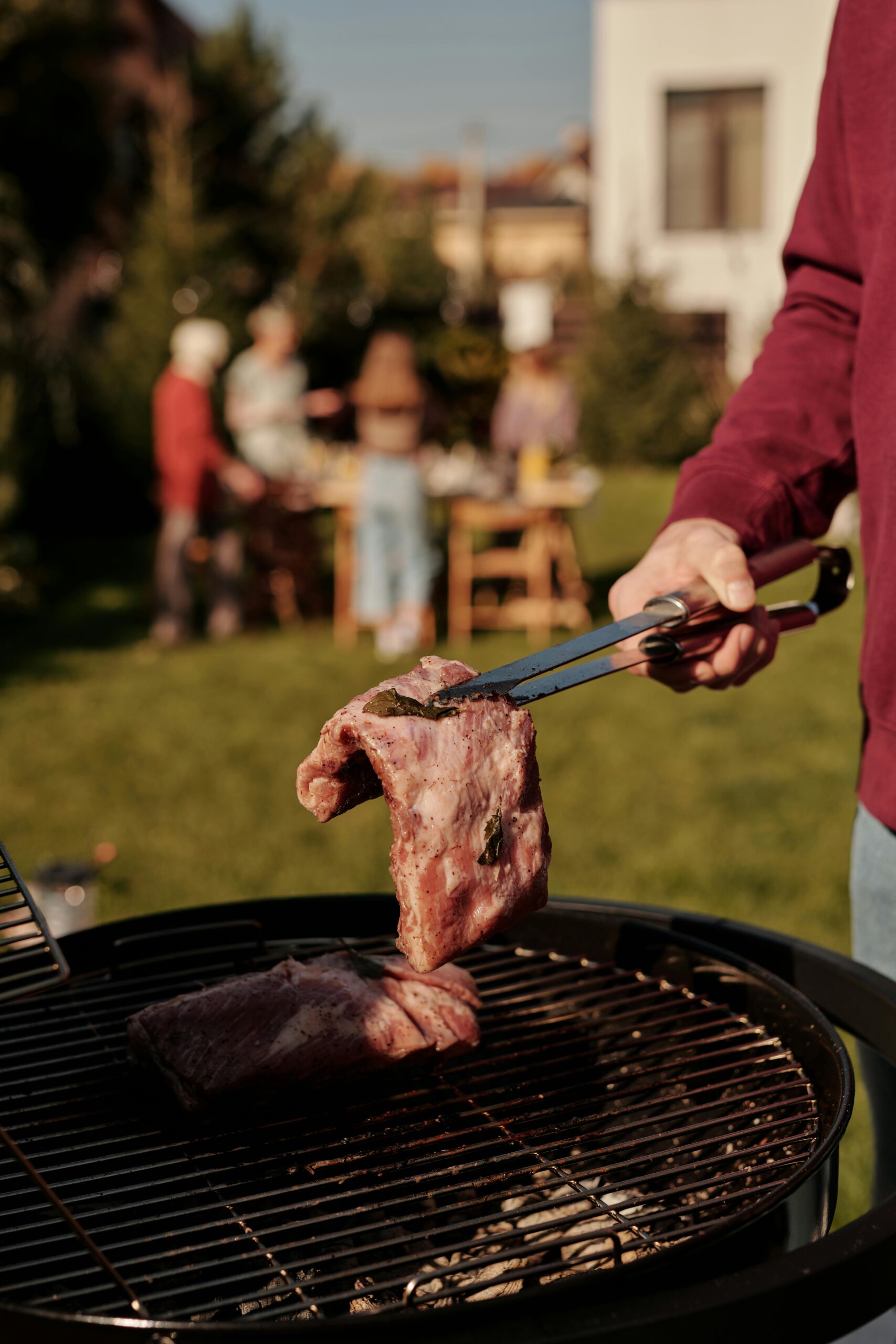 BBQ scene with grilling meat and people socializing in a sunny backyard setting.